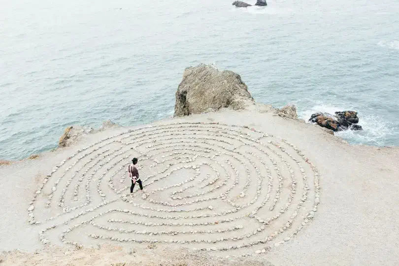 Personne marchant dans un labyrinthe de pierres disposées en cercles concentriques sur une plage près d'une falaise avec l'océan en arrière-plan.