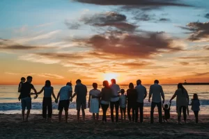 Groupe de personnes de différentes générations tenant les mains devant un coucher de soleil sur la plage, reflétant la synergie et la relaxation collective